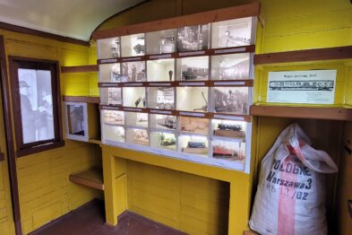 Wnętrze prezentuje żółty wagon pocztowy z epoki, wyposażony w drewniane szafki na listy i paczki. Na ścianie powieszono fotografie ilustrujące historię transportu kolejowego. W rogu widoczny jest work przewoźny, typowy dla poczty kolejowej.