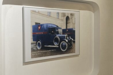W białej ramie znajduje się fotograficzne przedstawienie starego, niebieskiego samochodu pocztowego z napisem 
