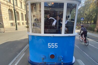 Niebiesko-biały tramwaj z numerem 555 stoi na torach ulicznych otoczony przez zabudowania miejskie. W tylnej części wagonu widać ludzi. Na asfalcie obok tramwaju porusza się rowerzysta.