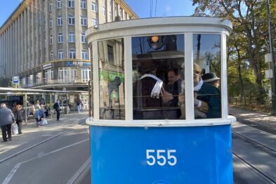 Historyczny tramwaj oznaczony numerem 555 stoi na torach w miejskiej przestrzeni, tłumy ludzi są w tle. Pojazd ma niebiesko-białe malowanie oraz otwarte okna, przez które widać pasażerów. W tle widać nowoczesne budynki i drzewa, jest słoneczny dzień.