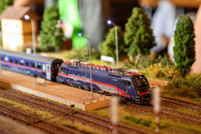 Model pociągu z niebiesko-czerwoną lokomotywą i pasażerskimi wagonami przejeżdża obok miniaturowego dworca kolejowego, otoczony realistycznymi drzewami i trawą. Sceneria makiety zawiera detale takie jak słupy oświetleniowe i miniaturowe znaki kolejowe, dodając realizmu ustawieniu. Tło rozmyte, co sugeruje głębię sceny i skupia uwagę na miniaturowym pociągu.