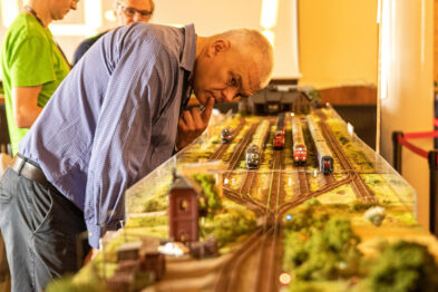 Osoba ogląda dużą makieta kolejową przedstawiającą tory, modele pociągów oraz krajobraz z drzewami i budynkami. Uwagę zwiedzającego skupiają szczegóły makiet, nad którymi się pochyla. W tle widać innych odwiedzających również zainteresowanych ekspozycją.