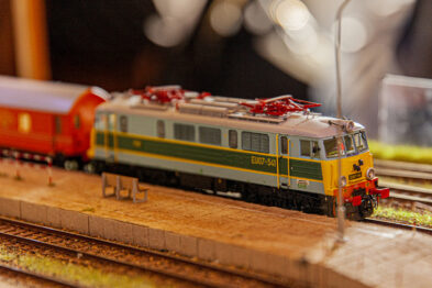 Model elektrycznego pociągu w skali H0 stoi na makiecie kolejowej, którą szczegółowo odwzorowano z prawdziwym krajobrazem, kompletnym z torami i otaczającą roślinnością. W tle widoczny jest czerwony wagon pasażerski, a całość jest oświetlona ciepłym światłem podkreślającym detale. Model jest koloru żółto-zielonego z czerwonymi elementami na przodzie lokomotywy i posiada pantografy umieszczone na dachu.