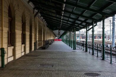 Pustkowiający peron dworca kolejowego z wiatą o metalowej konstrukcji wspartej na zielonych słupach. Podłoga peronu wyłożona jest kostką brukową i ciągnie się wzdłuż budynku dworcowego o jasnych ścianach. Na końcu peronu widać czerwony pociąg.