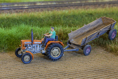 Model traktora z przyczepą stoi na makiecie pola, z wyraźnie zaznaczonymi śladami od kół. Osoba siedząca na traktorze jest skierowana w lewo. Makieta jest szczegółowa, z imitacją ziemi i trawy na szczegółowo odwzorowanym terenie.