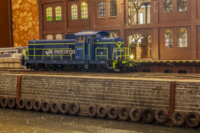 Model lokomotywy w kolorach niebiesko-żółtych stoi na torowisku obok ceglanej zabudowy, która imituje budynek przemysłowy z oknami, drzwiami i latarniami. Kolejowe wagony towarzyszą lokomotywie na sąsiednim torze. Całość wskazuje na szczegółowo opracowaną miniaturę kolejową, odzwierciedlającą typową scenerię stacyjną.