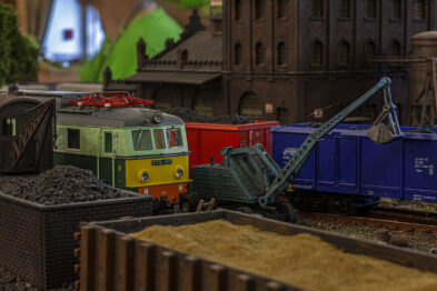 Model kolejowy przedstawia lokomotywę stojącą obok węglarki i koparki z układem linowym. W tle widać modele budynków przemysłowych i drzewa. Szczegółowość i kolory modeli wskazują na dbałość o realizm sceny.