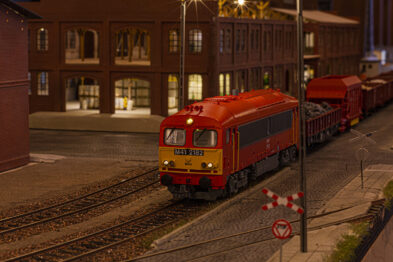 Czerwony model lokomotywy z serii H0 ciągnie za sobą zestaw czerwonych wagonów towarowych, poruszając się po makiety kolejowej, która odwzorowuje rzeczywiste otoczenie torów, w tym latarnie i budynki. Makieta zawiera szczegółowe elementy, jak torowisko z rozjazdami oraz sygnalizację kolejową, a tło tworzą wiernie odwzorowane budowle przemysłowe. Całe ujęcie jest oświetlone w sposób imitujący zmierzch