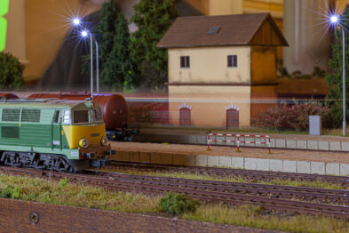 Model pociągu z zieloną lokomotywą i czerwonym wagonem stoi na makiecie kolejowej przedstawiającej peron i tory. Latarnie oświetlają scenę, imitując nocne oświetlenie. W tle widać modelowe budynki i drzewa, a całość utrzymana jest w skali H0.
