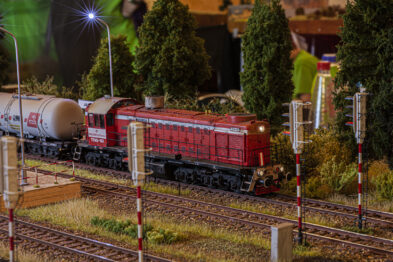 Model lokomotywy ciągnie za sobą szereg wagonów towarowych na makiety kolejowej. Torowisko przecina się z kilkoma innymi liniami, naszpikowane jest semaforami i latarniami. Tło stanowi imitacja zielonej roślinności i zabudowań.