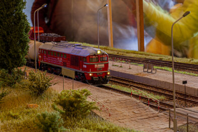 Model czerwonego lokomotyw spalinowego stoi na makiecie kolejowej, przedstawiającej perony i tory. Otoczenie makietowe jest szczegółowo dopracowane, z widoczną roślinnością i elementami małej architektury. W tle modelu widnieje nieostry obraz, który może być tłem scenicznym dla makiet kolejowych.