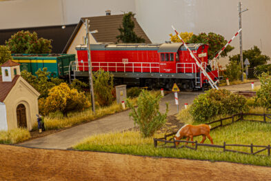 Model kolejowy przedstawia krajobraz z pociągiem przejeżdżającym przez przejazd kolejowy, podnoszące się zapory i sygnalizacja świetlna. Wokół rozpościera się zielony krajobraz z drzewami, pastwiskiem i figurką krowy, a w tle widoczny jest model budynku. Scena jest częścią większej makiety prezentującej sceny związane z kolejnictwem.