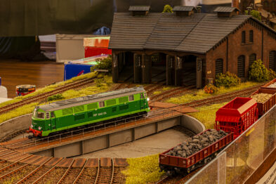 Model kolejowy przedstawia zielony lokomotywę elektryczną stojącą na obracalnej płycie segmentowej wewnątrz lokomotywowni, otoczony przez tory i modele pociągów towarowych z czerwonym wagonem i innymi elementami. Wokół są umieszczone miniaturowe modele budynków, roślinności oraz torowiska. Całość przedstawia scenę z makietą kolejową, oddającą atmosferę i szczegóły charakterystyczne dla stacji kolejowej.