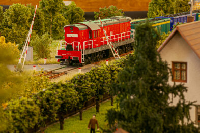 Czerwony model lokomotywy stoi na torach przy przejeździe kolejowym; otoczony jest przez drzewa i roślinność w różnych odcieniach zieleni. Z boku widoczny jest model domku z jasnożółtymi ścianami i ciemnoszarym dachem. Na pierwszym planie znajduje się figurka człowieka ubranego w ciemny strój, idącego obok wyższych roślin.