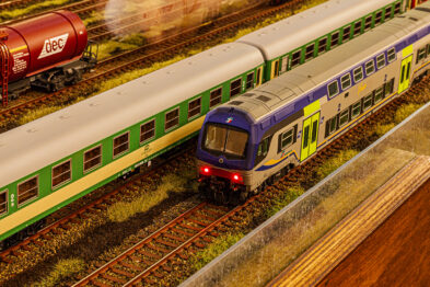 Model pociągu pasażerskiego ze szczegółowo odwzorowanymi wagonami i lokomotywą porusza się po miniaturowych torach kolejowych. Obok widoczne są inne modele kolejowe, w tym czerwony wagon towarowy. Całość prezentuje wysoki poziom detalu i realizmu, typowy dla makiety kolejowej skali H0.