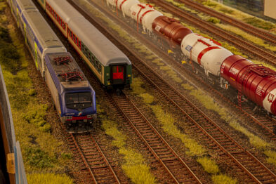 Model kolejowy przedstawia torowisko z rozmieszczonymi różnymi pociągami, w tym jeden pasażerski i kilka towarowych z cysternami. Szczegółowa makieta uwzględnia realistyczne detale takie jak trawa i podkłady kolejowe, a także modułowa konstrukcja torów. Makieta odzwierciedla rzeczywisty krajobraz kolejowy, ukazując starannie odwzorowaną infrastrukturę kolei.