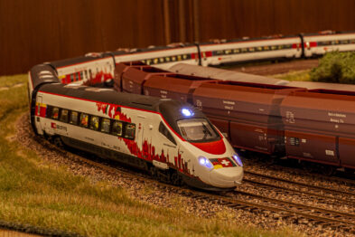 Model pociągu o białej i czerwonej kolorystyce z wyraźnym oświetleniem frontowym jeździ po makiecie kolejowej, przedstawiającej wielotorowe linie. Na drugim planie widoczne są wagony towarowe o brązowo-czerwonym zabarwieniu. Makietę zdobią wiernie odwzorowane detale takie jak tory, podkłady oraz otaczający krajobraz.