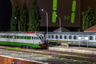 Model kolejowy przedstawia zielony elektrowóz stojący na torach obok dworca kolejowego; za modelem drzew wystają szczyty charakterystycznych budowli. W tle widoczne są modele budynków kolejowych oraz latarnie oświetlające scenę. Cyfrowy zegar w prawym górnym rogu ekspozycji wskazuje czas 3:22.