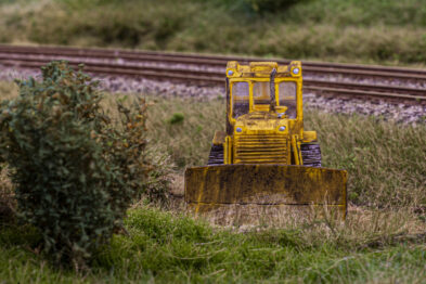 Miniaturka żółtej koparki stoi na trawiastym terenie obok modelu torów kolejowych, dająca iluzję faktycznych robót przy torach. Otoczenie jest szczegółowo wykonane z roślinnością, imitującą naturalne środowisko. Model zestawiony jest tak, aby wyglądał jak prawdziwy sprzęt w terenie.