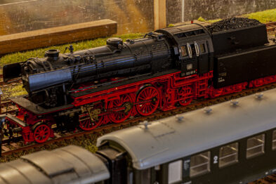 Makieta kolejowa przedstawia lokomotywę parową w kolorze czarnym z czerwonymi elementami w skali H0, stoi ona na torach obok innych wagonów. Dokładnie odwzorowany model zawiera wiele detali, takich jak koła, okna i schodki. Tło obrazu jest rozmazane, sugerując głębię pola i koncentrację na modelu pociągu.