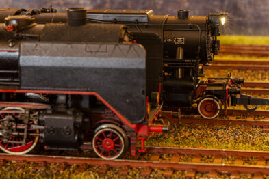Model parowozu stoi na makiecie kolejowej, z detalami takimi jak szyny i żwir. Parowóz ma czarny kolor z czerwonymi elementami na kołach, charakterystyczne dla lokomotyw parowych. Tło jest nieostre, co pozwala skupić się na modelu i jego szczegółach.