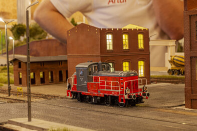 Model kolejowy czerwonej lokomotywy spalinowej stoi na torze przed budynkiem z cegły. W tle widoczny jest plakat z wielkoformatowym zdjęciem człowieka. Scena jest częścią makiety kolejowej, na której przeważają ciepłe, wieczorowe odcienie.