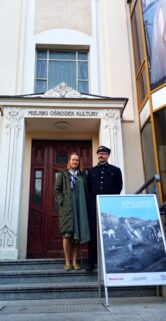 Kobieta i mężczyzna w historycznym mundurze kolejowym stoją przed wejściem do budynku z tablicą "MUZEUM KOLEJNICTWA", a obok nich znajduje się plakat z czarno-białym zdjęciem lokomotywy i ludzi. Na fasadzie budynku nad drzwiami widoczny jest napis "PAŁAC MŁODZIEŻY". Słońce oświetla scenę, a posadzka przed budynkiem wydaje się być utrzymana w czystości.