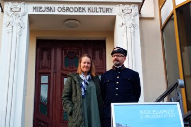 Kobieta i mężczyzna w historycznym mundurze kolejowym stoją przed wejściem do budynku z tablicą 