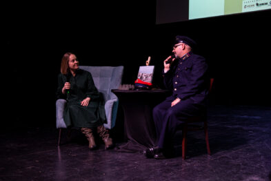 Dwoje ludzi siedzi na scenie; jeden w kolejarskim mundurze gestykuluje podczas rozmowy. Przed nimi stoi stolik z czerwonym obrusem, na którym leży książka i mikrofon. W tle znajdują się dwa fotele i projektor wyświetlający prezentację.