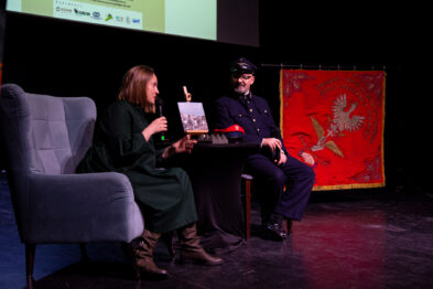 Kobieta i mężczyzna w historycznych strojach siedzą na scenie, mężczyzna ma na sobie mundur kolejarza. Kobieta trzyma kartkę i coś tłumaczy, a przed nimi stoi mikrofon na statywie. W tle widać czerwony sztandar z herbem.