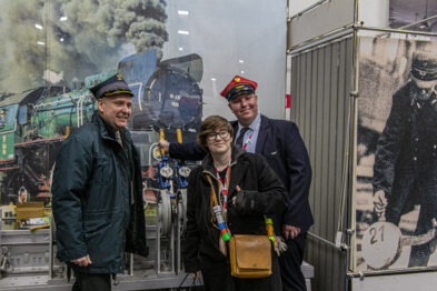 Trzy osoby w tradycyjnych kolejowych mundurach pozują do zdjęcia; dwóch mężczyzn i jedna kobieta trzymają się wzajemnie i uśmiechają. W tle znajduje się czarno-biała fotografia z motywem kolejowym, przedstawiająca parowóz i pracowników kolei. Scena odbywa się wewnątrz, potencjalnie w muzealnej czy wystawienniczej przestrzeni.