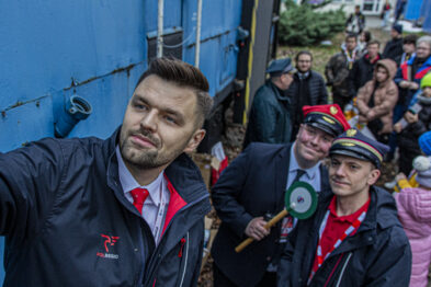 Grupa osób, w tym mężczyźni w kolejarskich strojach stoi przed niebieskim wagonem kolejowym; jeden z mężczyzn trzyma zielony sygnał dyskowy i wykonuje selfie. Tłum ludzi o różnym wieku siedzi na ławkach w tle, a wszyscy wydają się być skupieni na wydarzeniu. Człowiek robiący zdjęcie ma na sobie czarną kurtkę z czerwonym logotypem, a pozostali mężczyźni są ubrani w uniformy z daszkowymi czapkami.