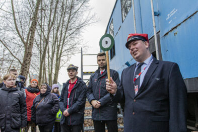 Grupa ludzi stoi obok niebieskiego wagonu kolejowego; jeden z mężczyzn, ubrany w czerwone nakrycie głowy i ciemny uniform, gestykuluje i wydaje się przemawiać do grupy. W tle widać drzewa bez liści i kolejny wagon, na którym widnieje okrągły znak z literą 