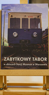 Książka o zabytkowym taborze kolejowym stoi na drewnianym stojaku. Okładka przedstawia pierwszy plan starego wagonu kolejowego z widocznym numerem EN57-1039. Tytuł na okładce głosi "ZABYTKOWY TABOR w zbiorach Stacji Muzeum w Warszawie".