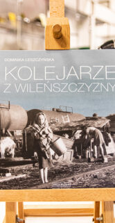 Na pierwszym planie stoi sztaluga z książką "Kolejarze z Wileńszczyzny". W tle dostrzec można modele kolejowe, w tym kolorowy parowóz i wagony. Oświetlenie zaakcentowuje detale ekspozycji i świadczy o dbałości muzeum o prezentowane eksponaty.