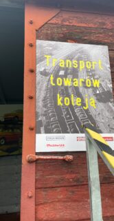 Plakat z napisem "Transport towarów koleją" jest wyeksponowany na drewnianej ścianie, przytwierdzony na czerwono-pomarańczowym tle z żółtym obramowaniem. Widoczne są detale konstrukcji, w tym metalowe wzmocnienie i śruby przymocowujące plakat. Na plakacie widać również torowisko oraz fragmenty żółtych elementów konstrukcyjnych sugerujących kolejowy kontekst.