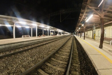 Peron kolejowy oświetlony w nocy, z ławkami i zadaszeniem, rozciąga się wzdłuż dwóch równoległych torów. Stacja wydaje się być pusta bez pasażerów czy pracowników. Na horyzoncie widać nadjeżdżający pociąg z włączonymi światłami przednimi.