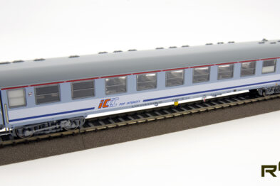 Model wagonu pasażerskiego o niebiesko-szarej kolorystyce stoi na makiecie torowiska; na boku wagonu jest logo przewoźnika kolejowego. Wagony posiadają szczegółowe oznakowania i wyraźnie widoczne są okna oraz drzwi. Model wygląda na starannie wykonany z wieloma realistycznymi detalami, odpowiadającymi rzeczywistemu pojazdowi kolejowemu.