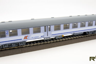 Model kolejowego wagonu pasażerskiego jest umieszczony na szynach, na białym tle. Wagon posiada niebiesko-szare barwy z napisem 