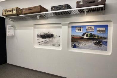 Przestrzeń wystawiennicza z fotografiami i modelami kolejowymi prezentuje pociągi w otoczeniu zimowym. Na ścianie wiszą dwa duże zdjęcia przedstawiające lokomotywy na śniegu, a nad nimi umieszczone są modele wagonów kolejowych. Całość tworzy eksponat tematyczny, oddający atmosferę pracy kolei w zimowych warunkach.