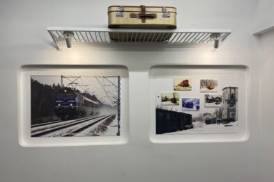 Na górnej półce umieszczony jest model lokomotywy nad dwoma dużymi fotografiami na białej ścianie. Lewa fotografia przedstawia elektryczną lokomotywę na torach kolejowych okrytych śniegiem. Prawa fotografia wyświetla wnętrze budynku kolejowego z różnymi przedmiotami, w tym taboretami i regałem z narzędziami.