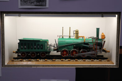 Model XIX-wiecznej amerykańskiej lokomotywy parowej i wagonu jest umieszczony w szklanej gablocie. Lokomotywa jest koloru zielonego z czerwonymi elementami, a obok niej stoi zielony wagon osobowy. Model umieszczony jest na makiecie torów, a w tle widnieje neutralna, jednobarwna ściana.