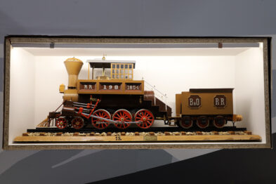 Model parowej lokomotywy z XIX wieku i dwóch wagonów jest eksponowany w szklanej gablocie. Lokomotywa ma duże, czerwone koła oraz klasyczne dla tamtego okresu ozdobne elementy. Na bocznej stronie wagonu widnieje napis 