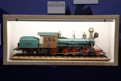 Model XIX-wiecznej amerykańskiej lokomotywy parowej jest prezentowany w białej gablocie. Lokomotywa ma zielone i brązowe elementy, a także numery i ozdoby. Obok lokomotywy dostrzegalne są również detale takie jak latarnie czy koła.
