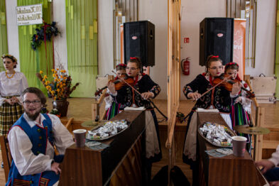 Trójka dzieci w tradycyjnych strojach gra na skrzypcach, siedząc przy drewnianych stolikach rozstawionych w pomieszczeniu z widocznymi głośnikami. Za nimi stoi kobieta w regionalnym ubiorze, a całe pomieszczenie wydaje się być przestrzenią, w której odbywa się wydarzenie kulturalne. Na stole przed dziećmi leżą opakowane słodycze, które mogą być częścią lokalnego zwyczaju lub upominkami dla uczestników.