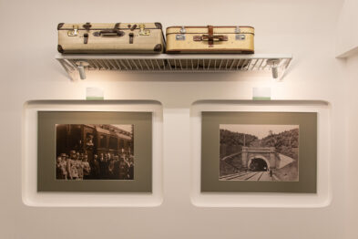 Dwa czarno-białe zdjęcia o tematyce kolejowej są eksponowane na jasnej ścianie, każde w białej ramie. Nad nimi zamontowana jest półka, na której ustawiono dwa stylowe walizki, sugerujące podróż koleją. Całość tworzy harmonijny układ, który podkreśla związek podróży z historią kolei.
