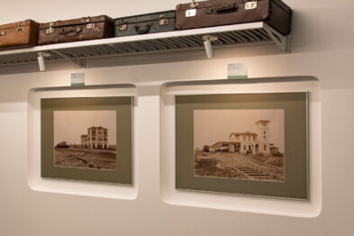 Na ścianie zawieszone są dwa oprawione fotografie przedstawiające budynki kolejowe w starym stylu. Powyżej fotografii umieszczono na półce kilka starych walizek. Oświetlenie akcentuje każdą z fotografii, a całość sprawia wrażenie starannie zaaranżowanej ekspozycji tematycznej.