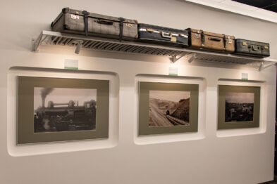 Ściana galerii pokazuje cztery czarno-białe fotografie o tematyce kolejowej oprawione w jednolite ramki. Nad zdjęciami znajduje się model pociągu towarowego umieszczony na półce. Każda fotografia przedstawia inne aspekty związane z koleją, od lokomotyw po sceny z życia stacji.