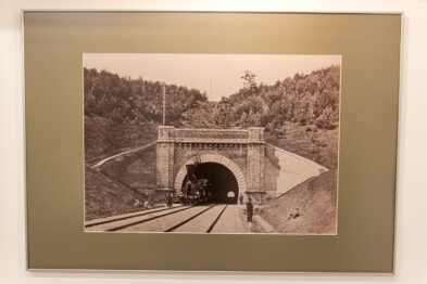 Czarno-biała fotografia przedstawia parowóz wjeżdżający do kamiennego tunelu kolejowego. Symetrycznie ułożone tory prowadzą wzrok w stronę otwartej konstrukcji tunelu. Zdjęcie umieszczone jest w jasnej oprawie na ścianie, co sugeruje wystawę prac fotograficznych.