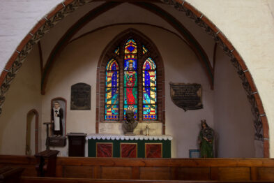 Wnętrze kościoła z widoczną kolorową witrażową okiennicą, po obu stronach których umieszczono pamiątkowe tablice. Na jednej z nich widnieje napis w języku polskim. Po bokach okna stoją dwie figury postaci, jedna przypomina zakonnika, a druga ubrana jest w zielone szaty z atrybutami koła i miecza.